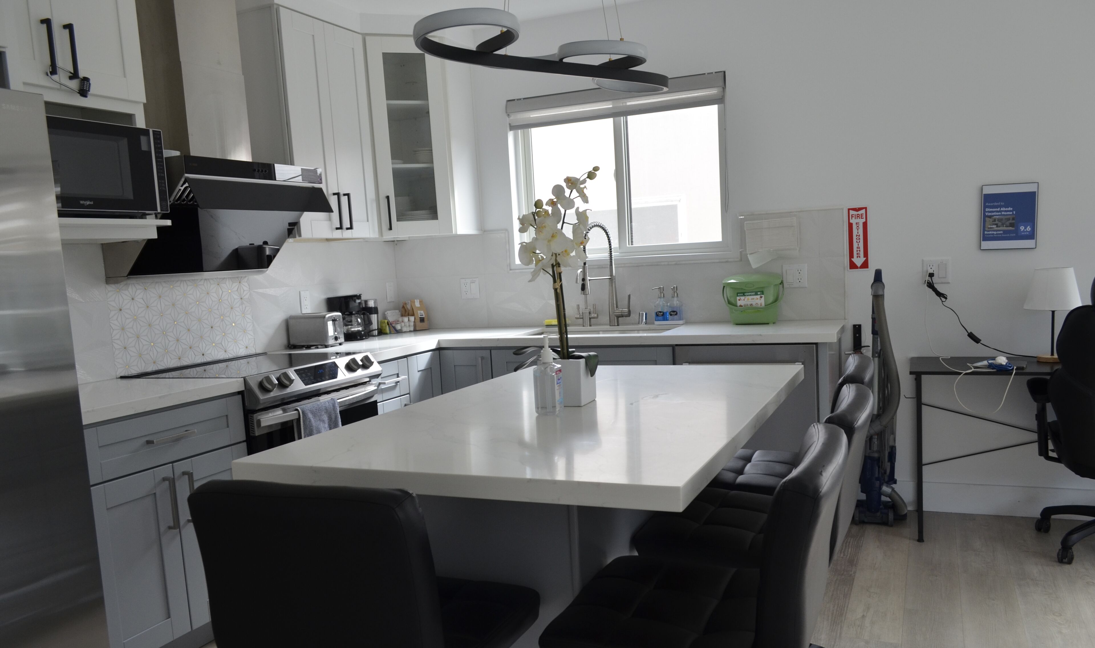 A modern kitchen with white cabinets, a built-in microwave, a coffee maker, and a sink. There is a window providing natural light and a small office area with a desk and chair.