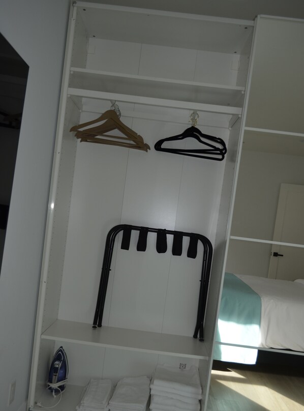 Ein weißer Kleiderschrank mit Kleiderstange, ein Bett mit türkisfarbenem Überwurf und ein blaues Bügeleisen auf dem Boden.