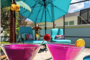 Deux verres à martini contenant un liquide rose et décorés de quelques éléments, un parasol bleu et une scène se déroulant au bord d’une piscine, avec une chaise et un palmier.