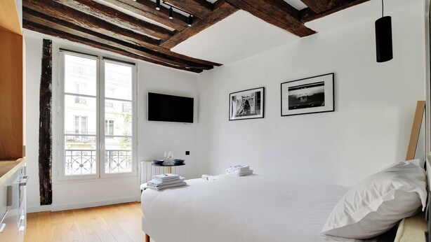 A hotel room with a large bed, a flat-screen TV, two framed pictures on the wall, and a balcony with a view of buildings. - Charming studio A/C - 2P - Le Marais (Paris)