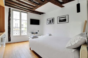 A hotel room with a large bed, a flat-screen TV, two framed pictures on the wall, and a balcony with a view of buildings.