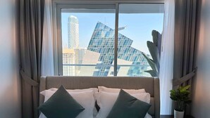 A hotel room with a large window, a bed with white linens and two dark green pillows, a small potted plant, and a view of modern buildings.
