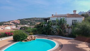 Una piscina con bordo curvo, circondata da un vialetto in mattoni e rigogliosa vegetazione.