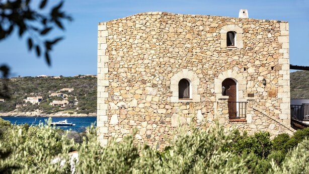Un edificio in pietra con finestre ad arco e un balcone, affacciato su una zona costiera con case e barche.