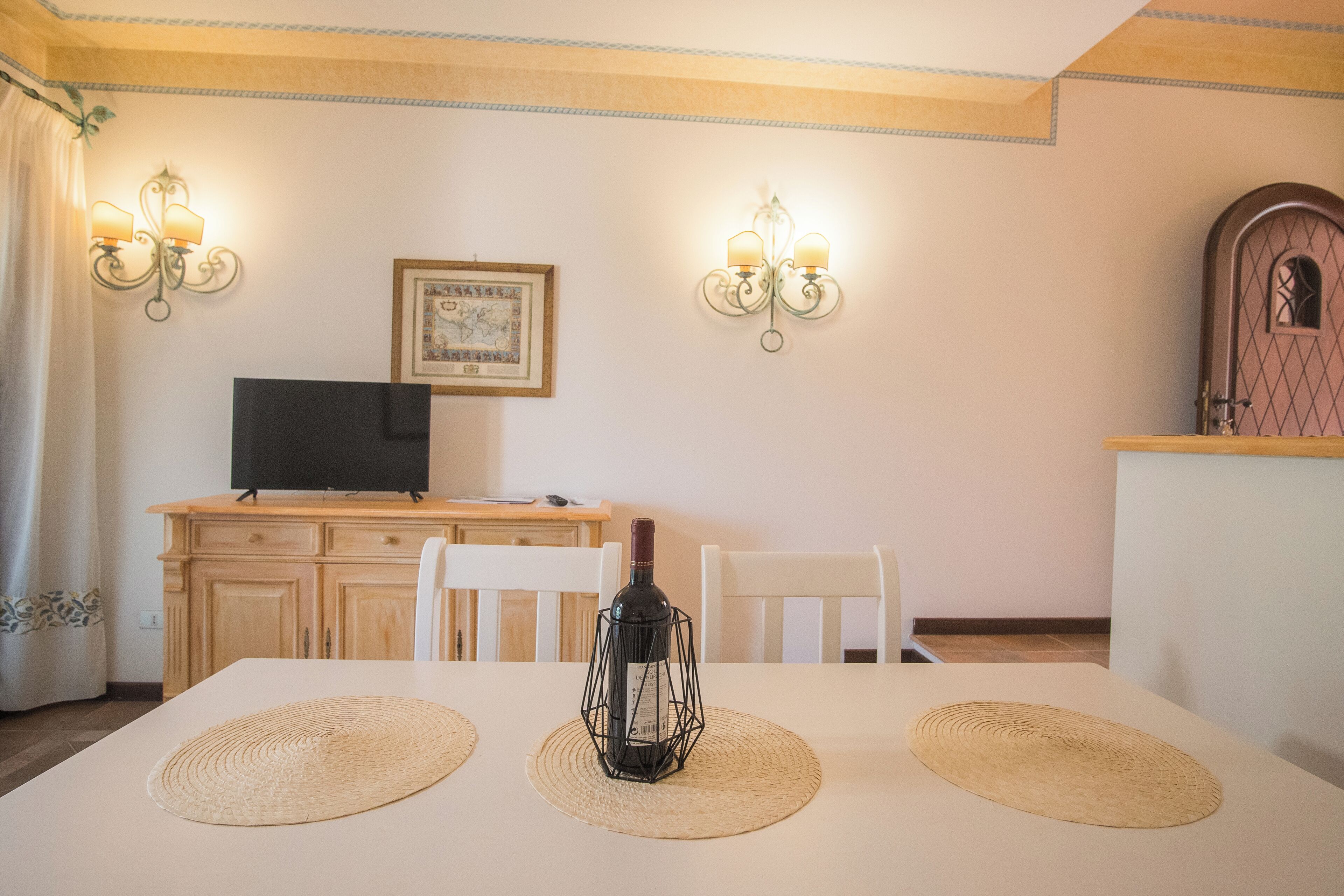A dining area with a table set for two, two wall-mounted light fixtures, a television on a wooden cabinet, and a framed picture on the wall.