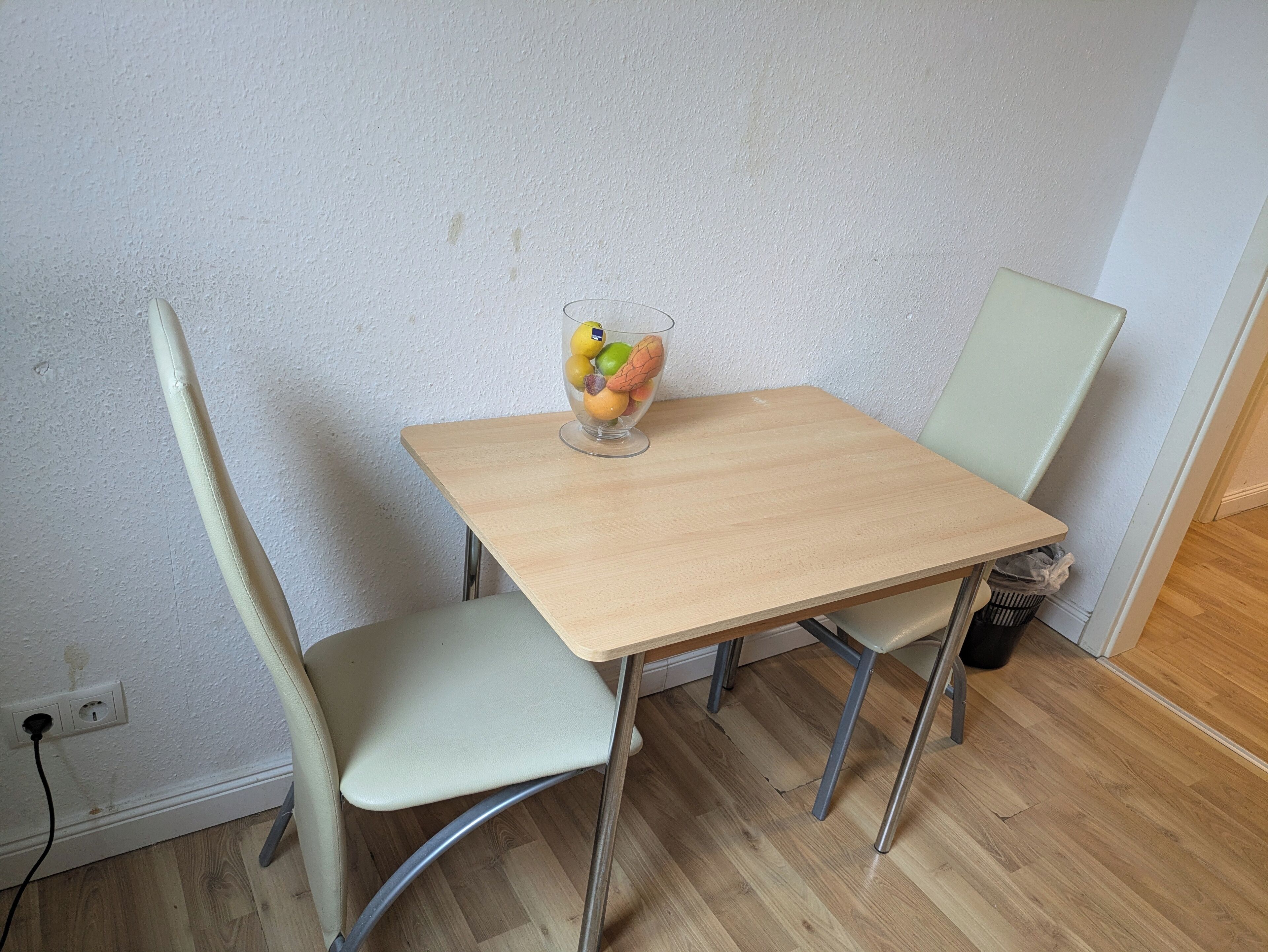A small wooden table with a glass bowl of fruit, a light-colored chair, and a plain wall with a power outlet.