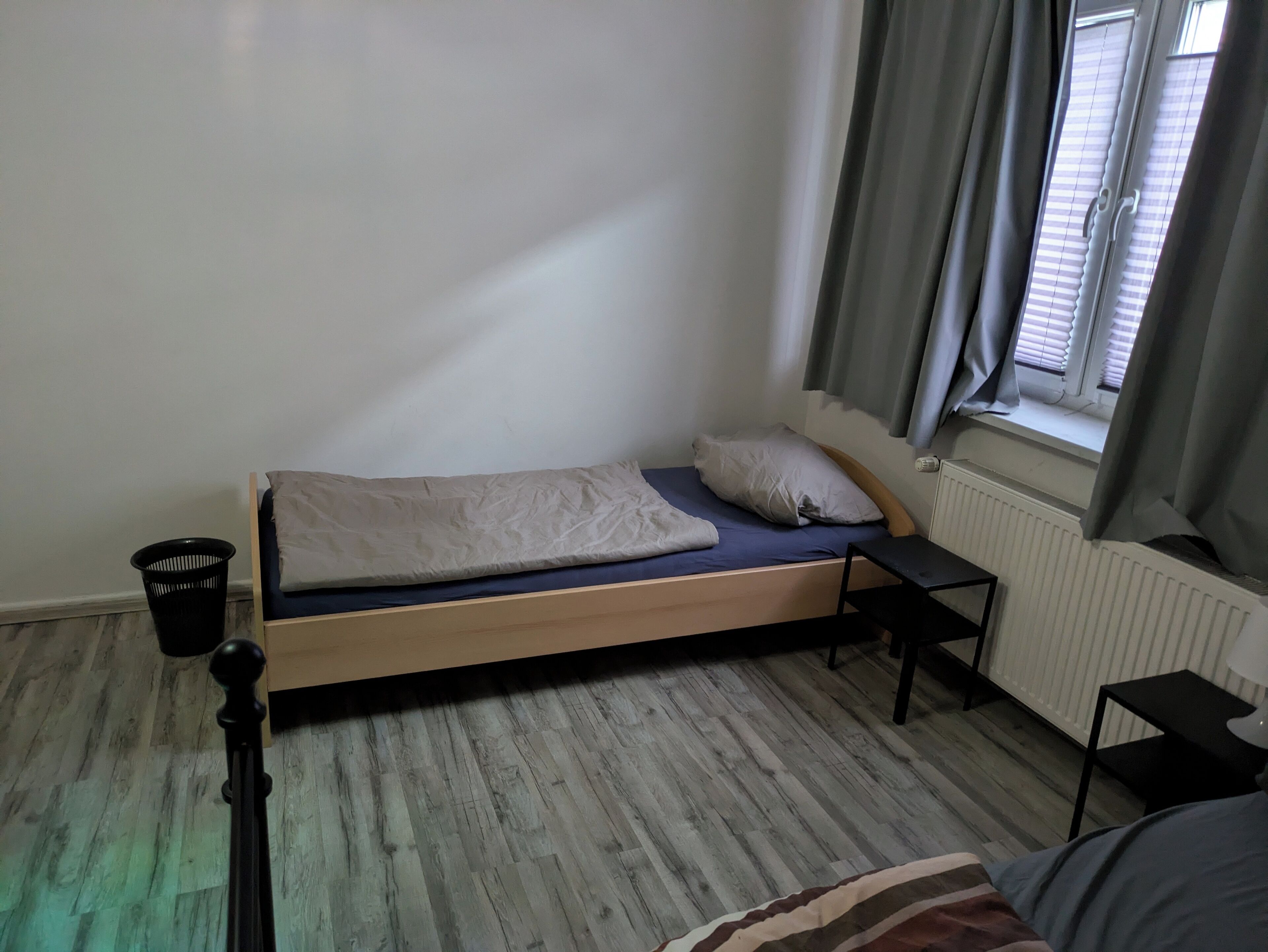 Une petite chambre avec un lit simple, un cadre en bois, un matelas bleu et une couverture blanche. On y trouve un présentoir en métal noir, une poubelle noire et une fenêtre avec des rideaux gris.