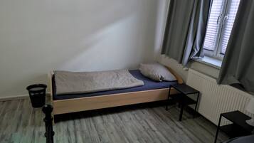 Une petite chambre avec un lit simple, un cadre en bois, un matelas bleu et une couverture blanche. On y trouve un présentoir en métal noir, une poubelle noire et une fenêtre avec des rideaux gris.