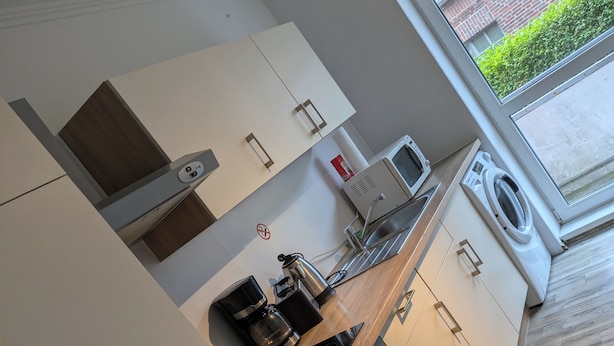 A modern kitchen with white cabinetry, a built-in oven, a coffee maker, and a window overlooking a garden. - NestOaseWHV Zimmer zw.City und Südstrand (Wilhelmshaven)