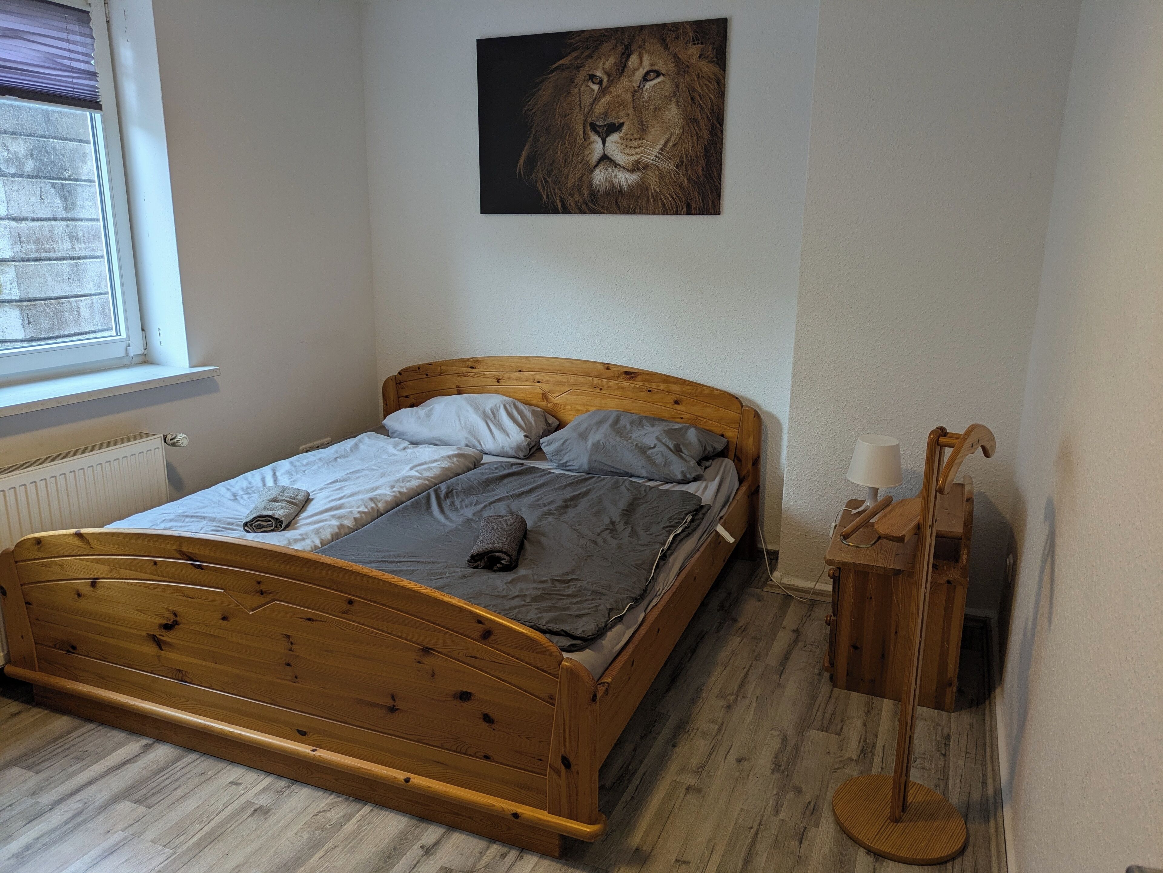 Uma cama de madeira com roupa de cama cinza, um radiador, uma janela com persianas e uma mesa de cabeceira de madeira com uma lâmpada.