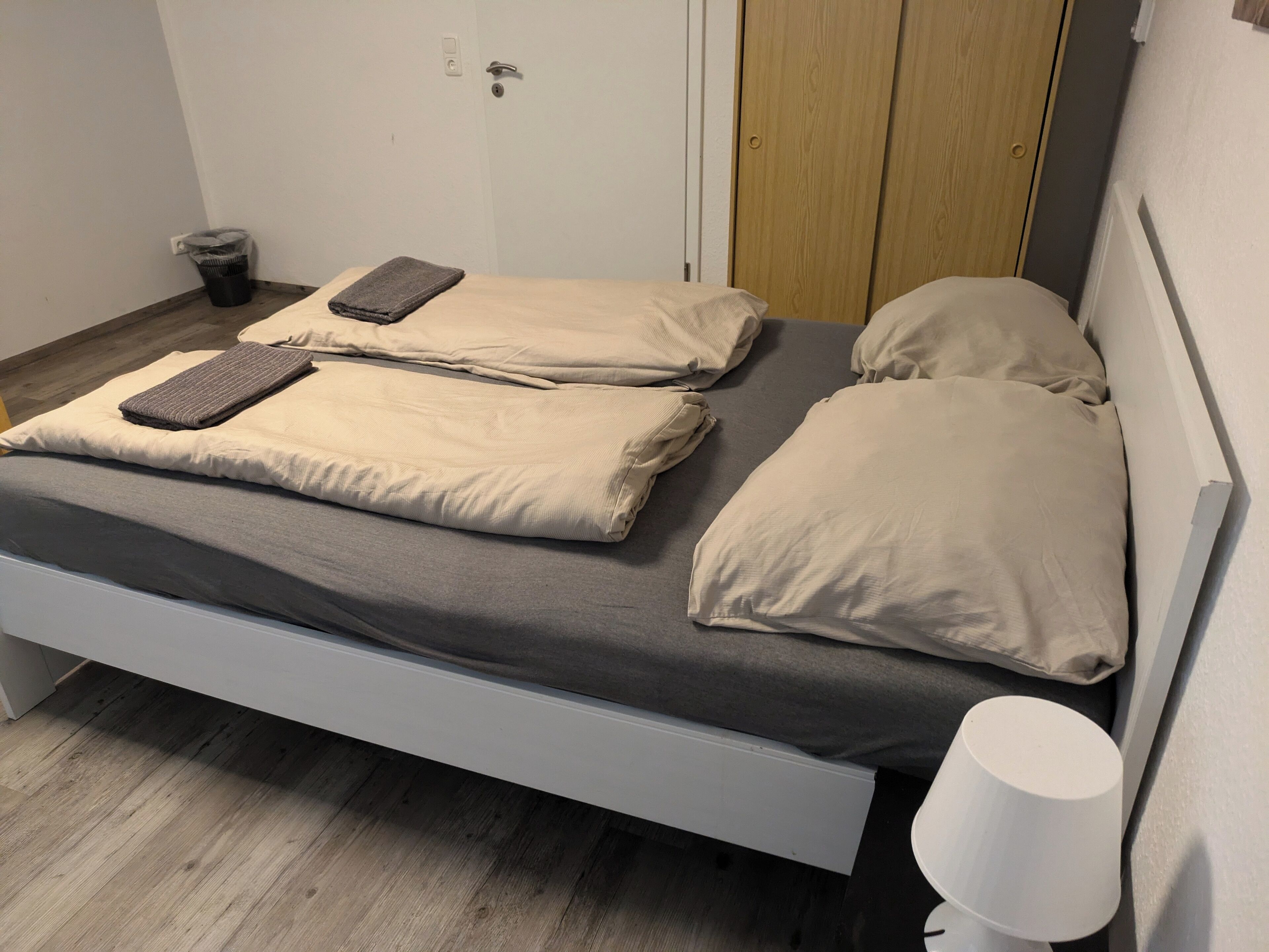 A single bed with a grey mattress and beige pillows, a bedside table with a lamp, and a wooden door in the background.