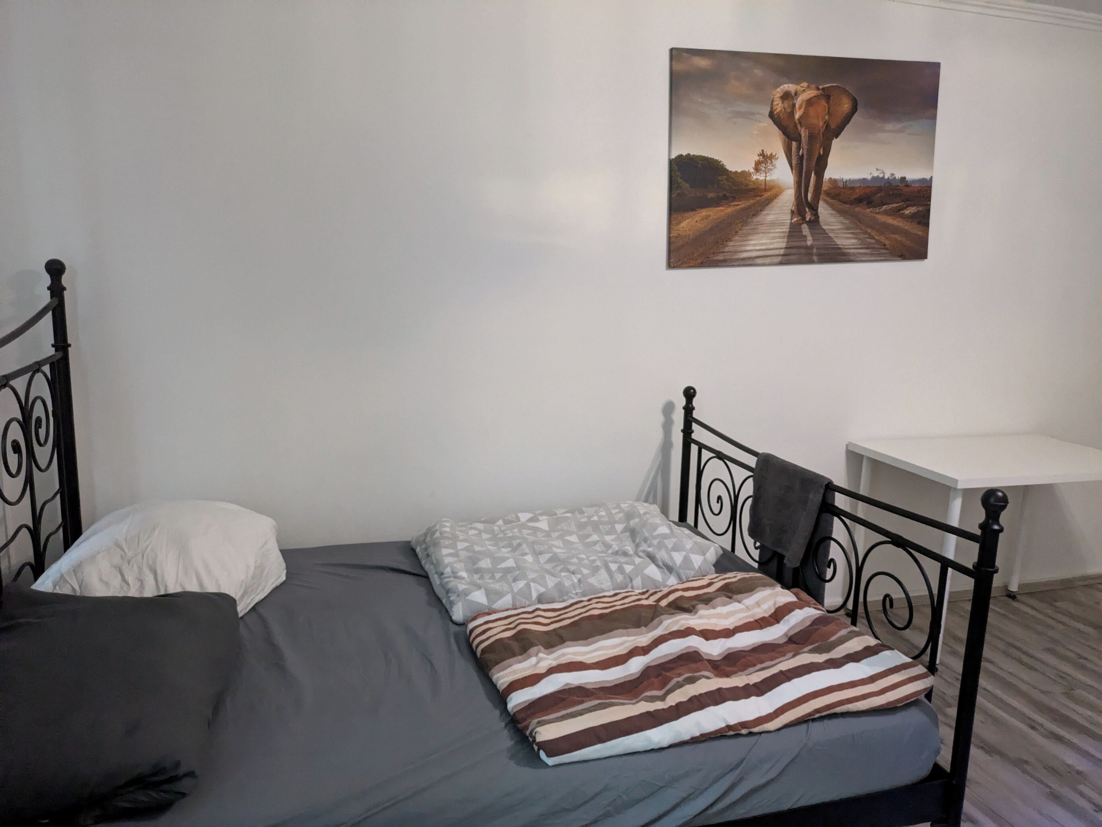 Un lit simple avec un matelas gris, un oreiller rayé et un oreiller blanc. Un tableau représentant un éléphant sur une route est accroché au mur.