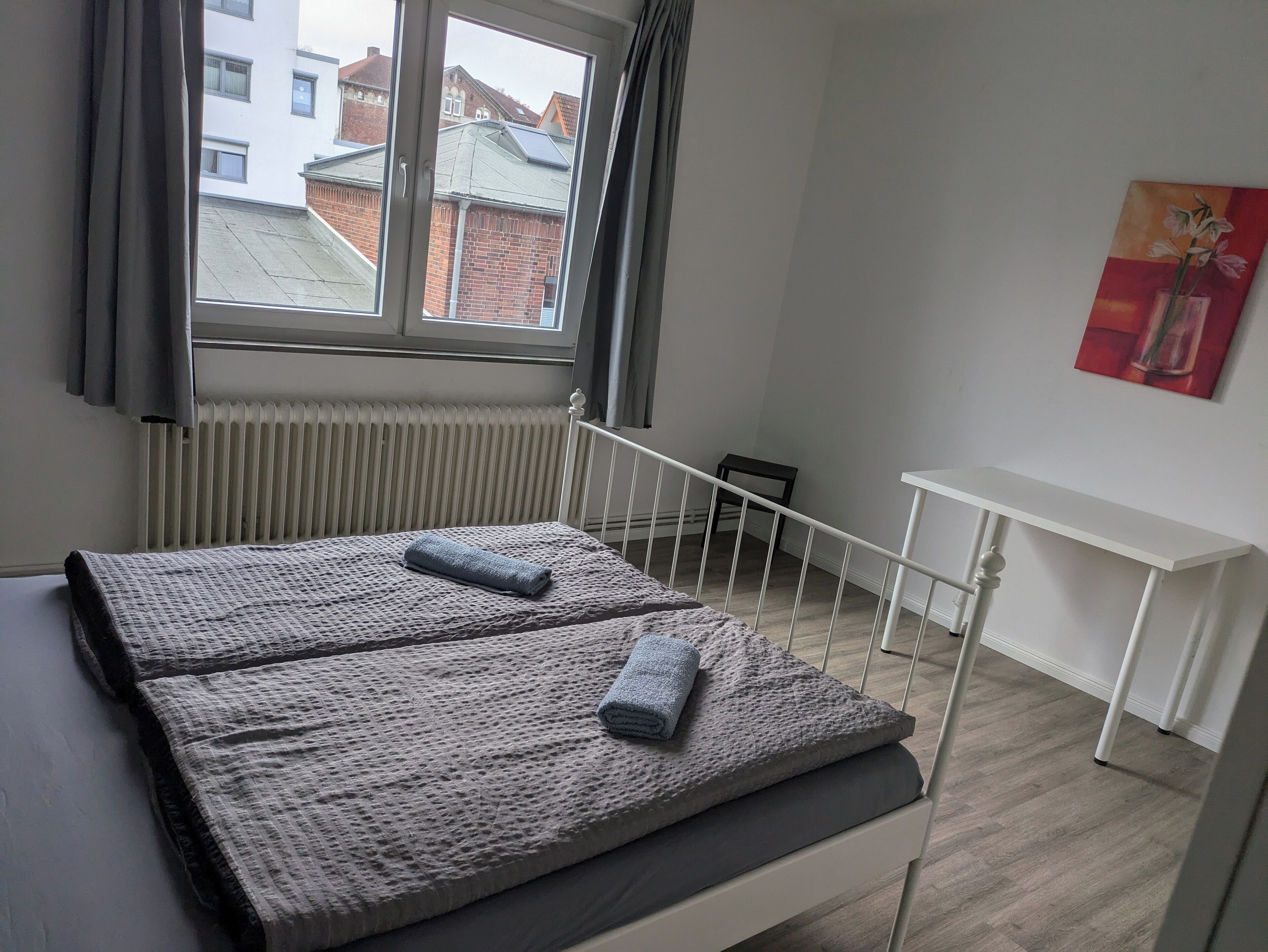 Un dormitorio con cama, ventana, radiador, una mesita de noche blanca y un cuadro en la pared.