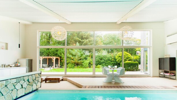 A modern indoor pool area with a stone wall, a dining table with chairs, and large windows offering a view of a lush garden. - Lanaken Retreat Pool Jacuzzi and Sauna (Lanaken)