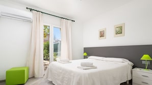 A modern bedroom with a large bed, two bedside tables, a green ottoman, and a view of greenery outside. - PLAYMAR (Alcúdia)