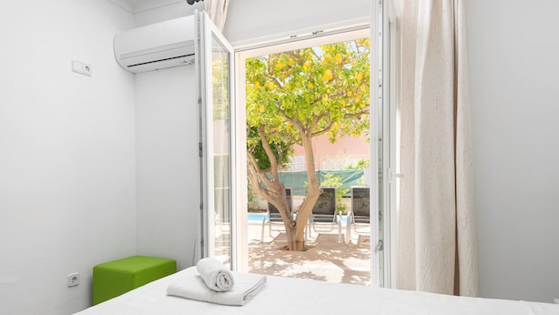 A hotel room with a bed, a green stool, and a view of a tree and pool outside. - PLAYMAR (Alcúdia)