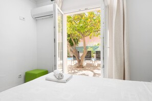 A hotel room with a bed, a green stool, and a view of a tree and pool outside. - PLAYMAR (Alcúdia)