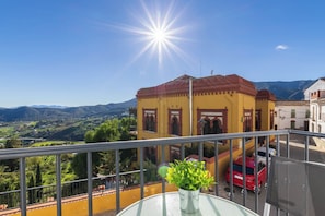 A balcony with a view of a yellow building, a red car, and a mountainous landscape.