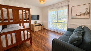 A room with a wooden bunk bed, a grey sofa, a television, and a window with a view of greenery.
