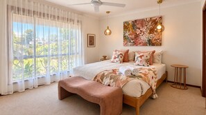 A bedroom with a large bed, a bench, a ceiling fan, a wall-mounted light, and a window with sheer curtains. - Broadwater Break: Stylish Coastal Retreat for Families (Broadwater)