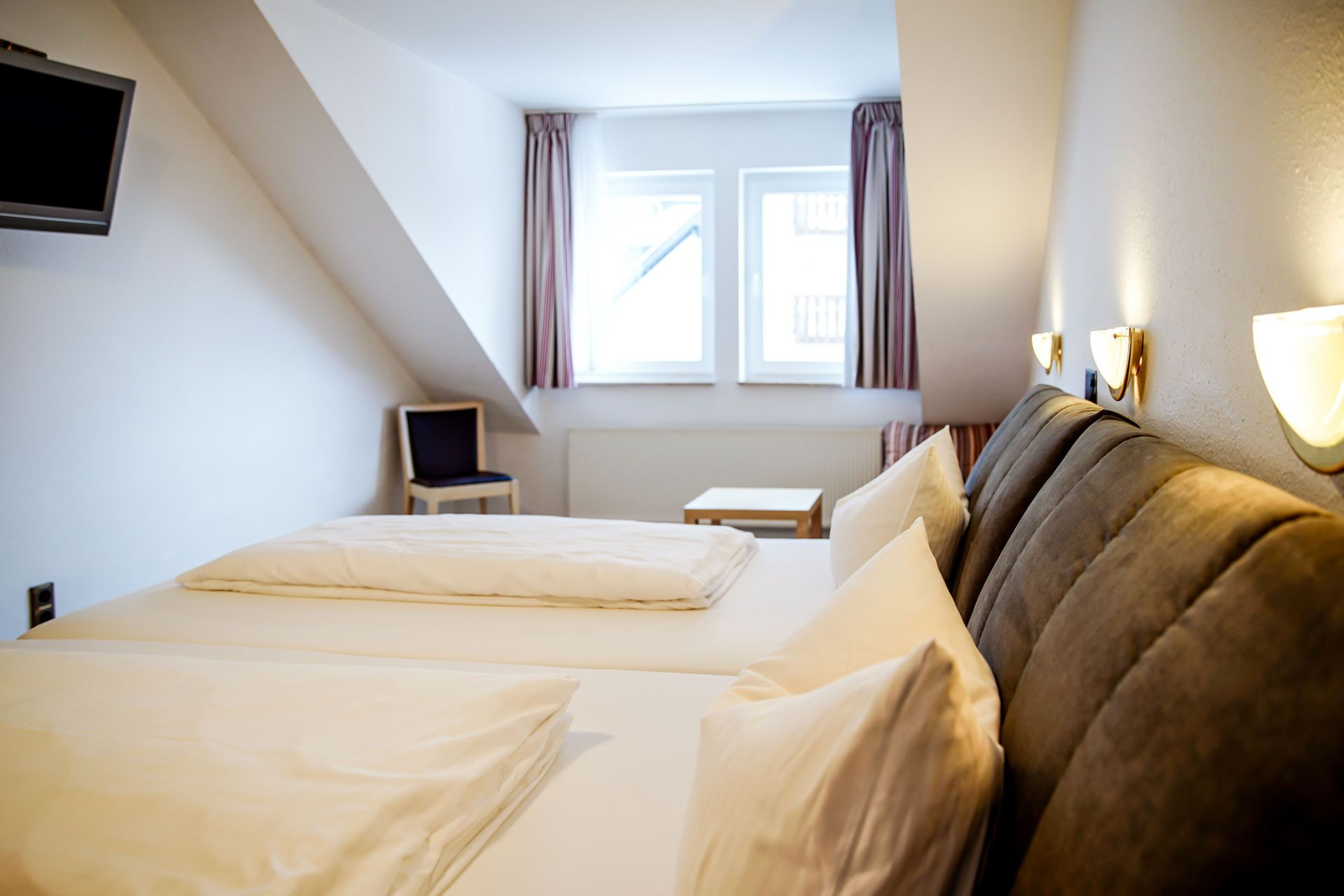 Et hotelværelse med to senge, en stol, et lille bord og et vindue med gardiner.
