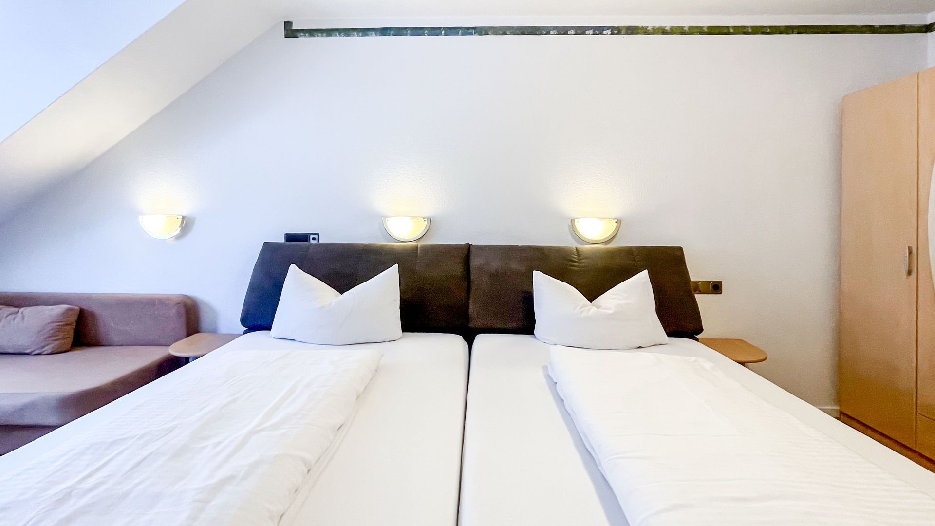 A double bed with white bedding and two pillows, a headboard with wall-mounted lights, and a wooden wardrobe.