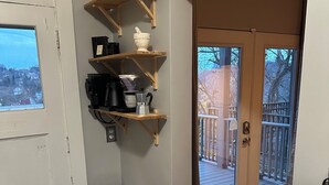 A compact kitchen with a gas stove, a trash bin, and wooden shelves holding kitchen items. A sliding glass door leads to an outdoor deck. - Pittsburgh city views close to all, cozy & bright 1BR - King + Dedicated Office (Pittsburgh)