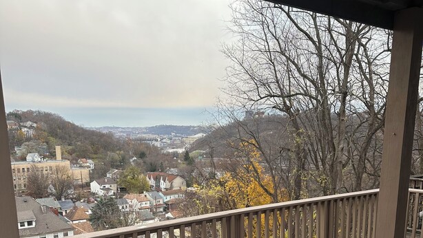 A covered outdoor deck with a view of a town, featuring a railing, a table, and a colorful rug. - Pittsburgh city views close to all, cozy & bright 1BR - King + Dedicated Office (Pittsburgh)