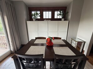 Un comedor con una mesa, sillas, un florero rojo y una ventana con cortinas.