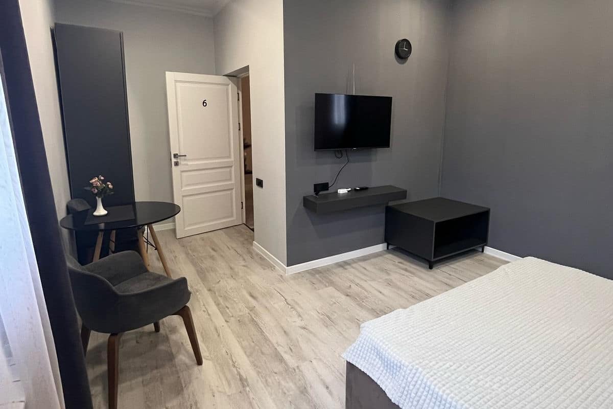 A modern bedroom with a bed, a TV mounted on the wall, a small table with a chair, and a door leading to another room.