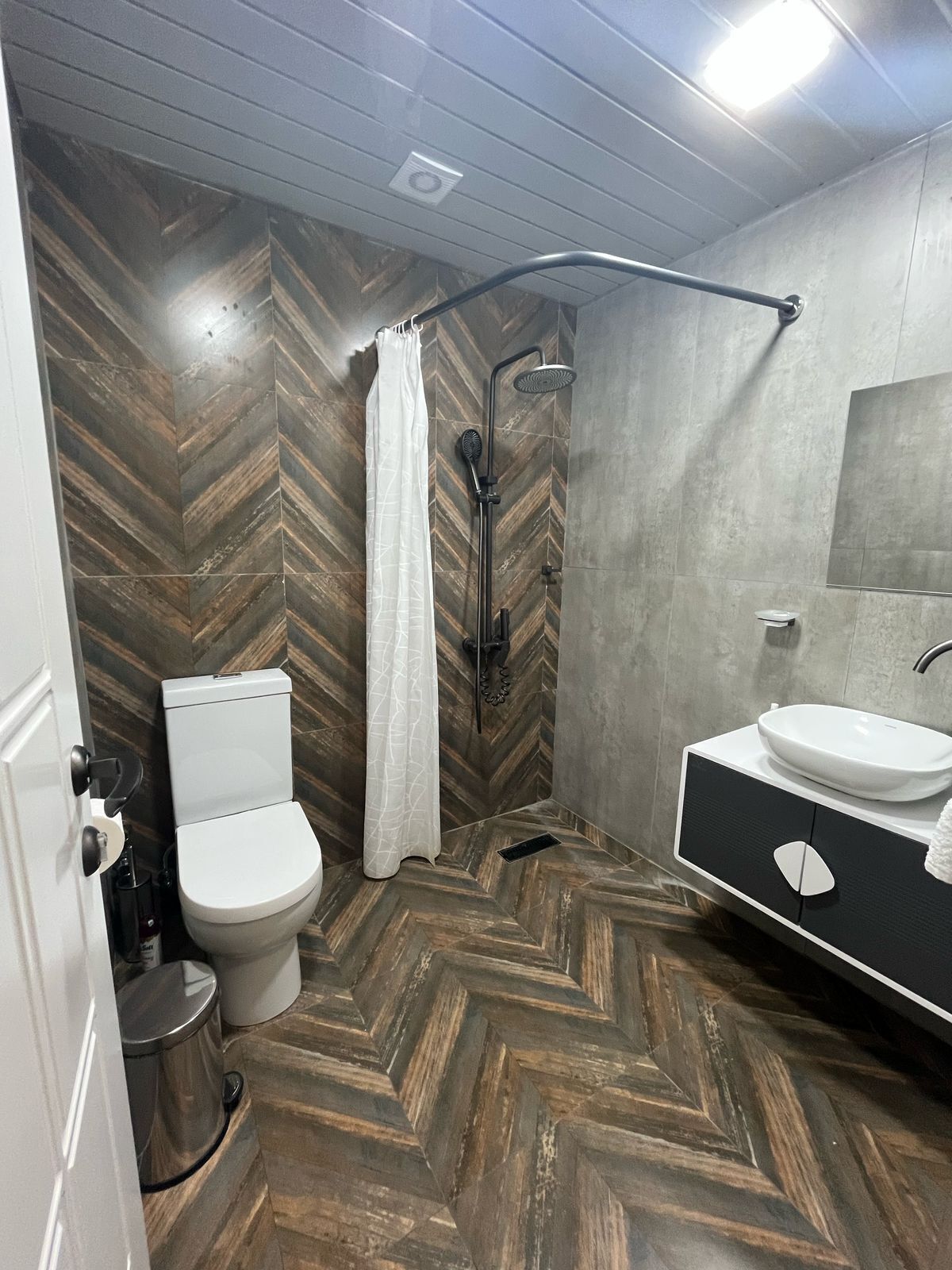 A modern bathroom with a herringbone wood floor, a white toilet, a shower with a curved design, and a black and white vanity with a mirror.