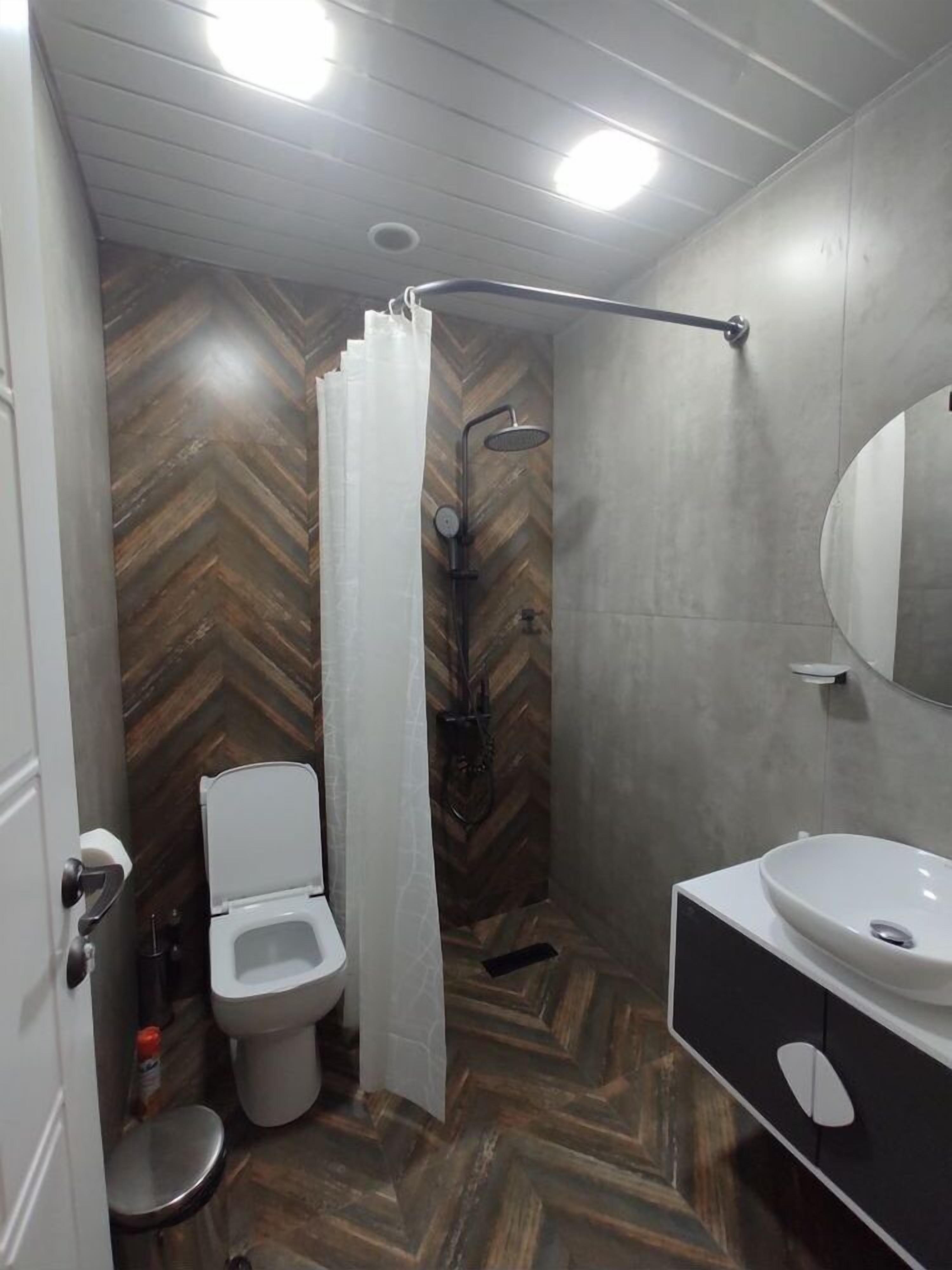 A modern bathroom with a herringbone wood floor, a shower with a curved showerhead, a toilet, and a vanity with a mirror.