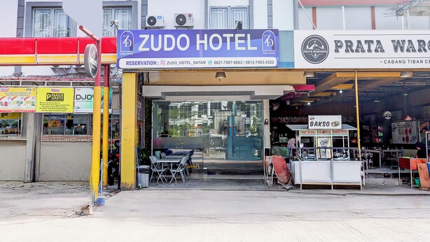 A ZUDO Hotel with a glass entrance, a visible outdoor seating area, and a sign for a nearby PRATA WARGO store.