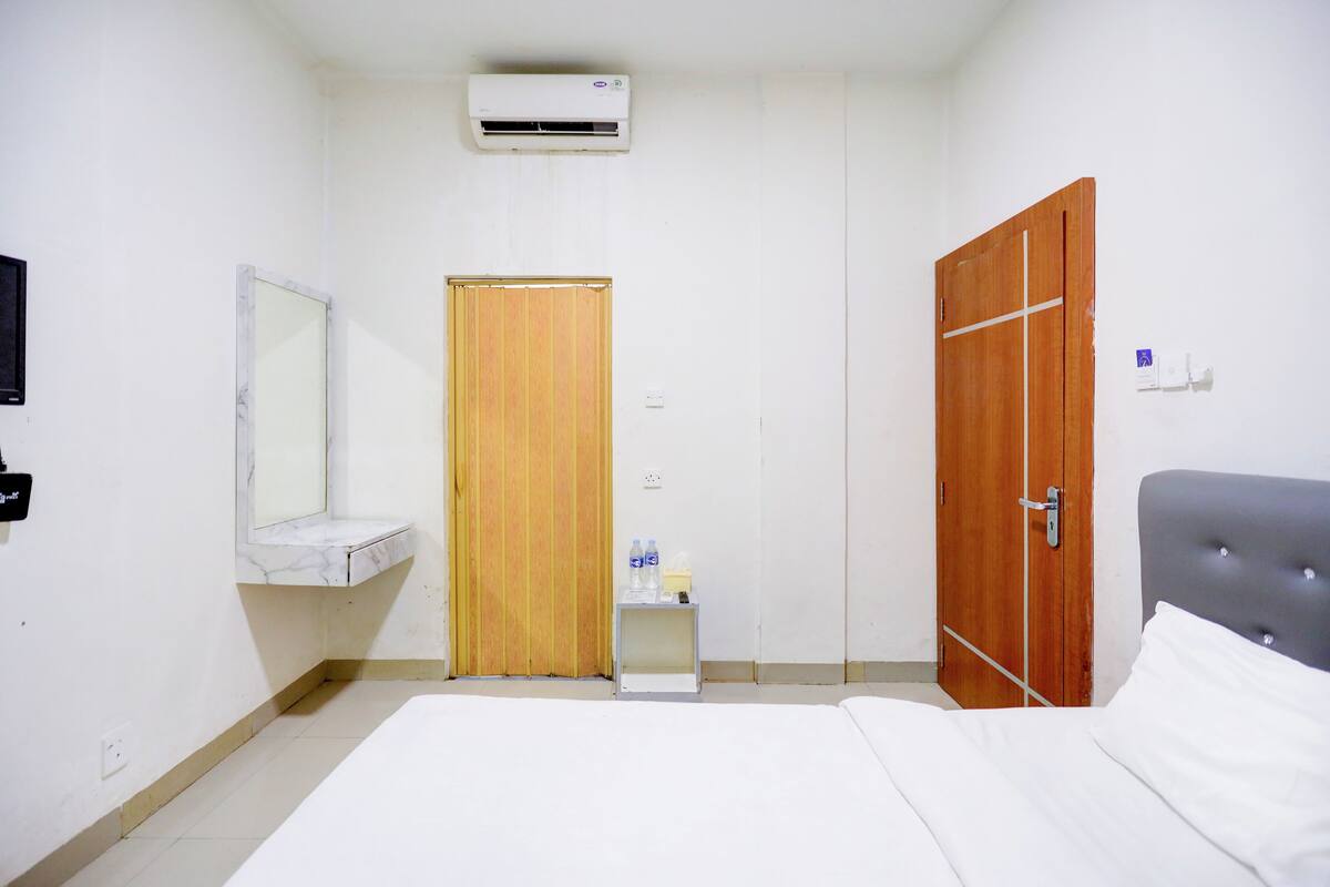 A hotel room with a bed, a wooden wardrobe, a small bedside table with a mirror, and an air conditioning unit on the wall.