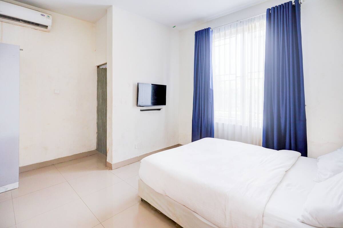 A hotel room with a bed, a television, a window with blue curtains, and an air conditioning unit.
