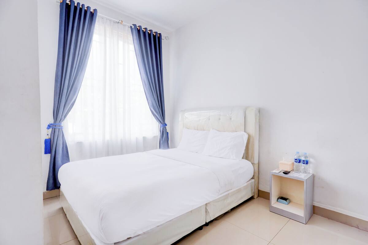 A neatly made bed with white linens, a bedside table with a tissue box and bottled water, and a window with blue curtains.