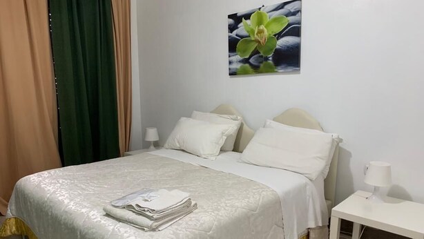 A neatly made bed with a white and yellow bedspread, two white pillows, and a folded white towel on the bed. There is a white nightstand with a lamp beside the bed. A green curtain is partially visible on the left side of the image. - Deluxe Room – ALH Hotel Digitale (Taranto)