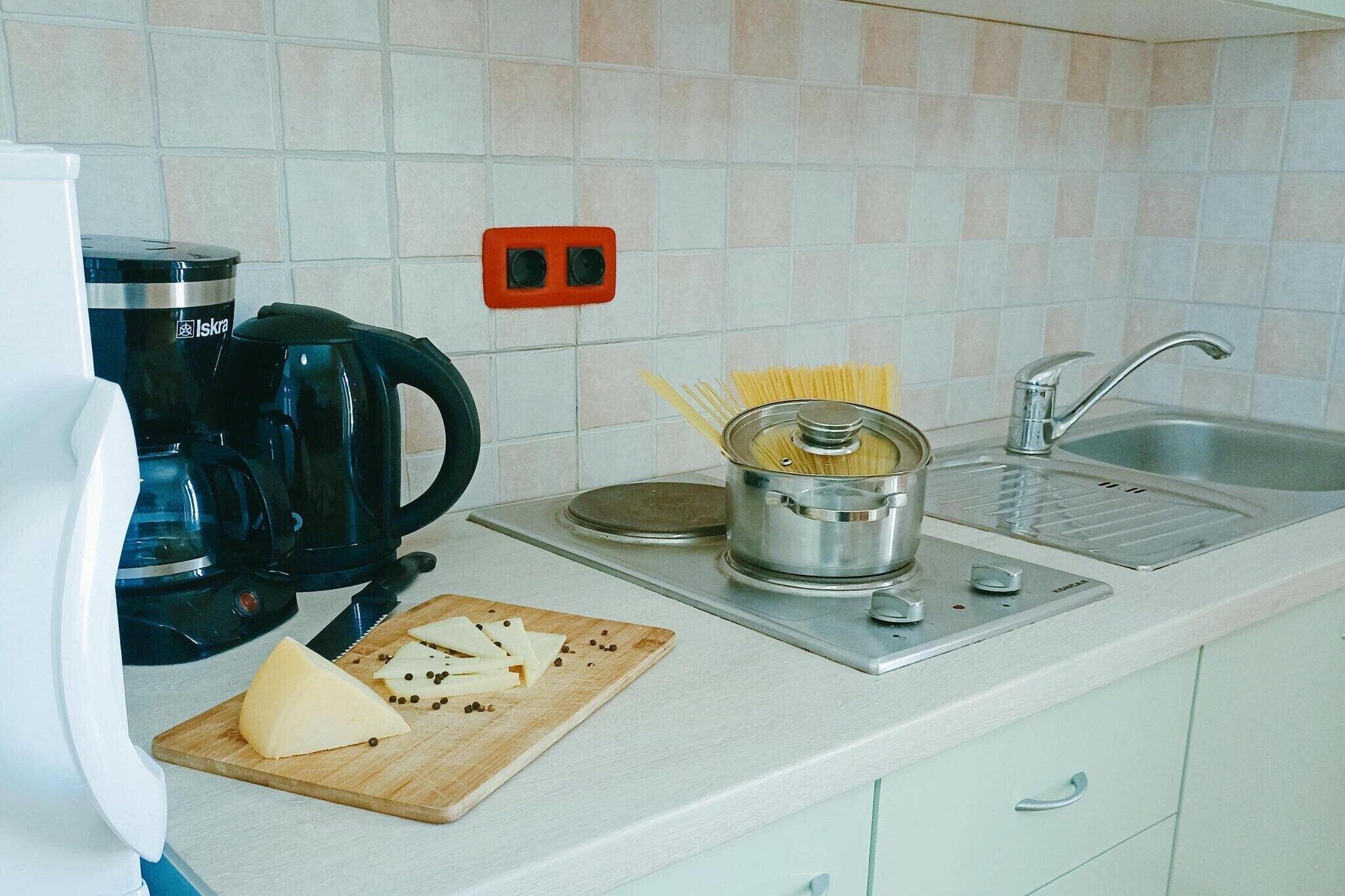 A kitchen with a coffee maker, a pot of spaghetti, a cutting board with cheese, and a sink.