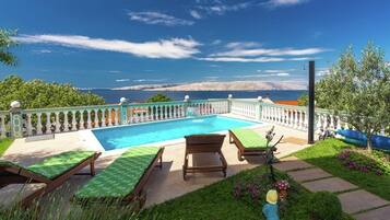 A pool with a view of the sea, surrounded by a garden with a fence and lounge chairs.