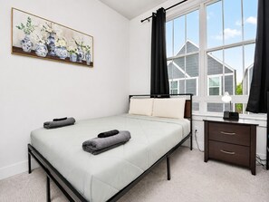 Una camera da letto con un letto, un comodino, una scrivania, una finestra con tende e un quadro appeso al muro.