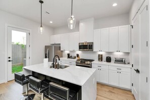 A modern kitchen with white cabinetry, a central island with bar stools, stainless steel appliances, and pendant lighting. - Central Houston Stay – 4BR Retreat by Downtown (Houston)