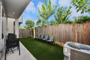 Un giardino sul retro con erba sintetica, un barbecue, una ventola e una fila di sedie addossate a un recinto in legno.