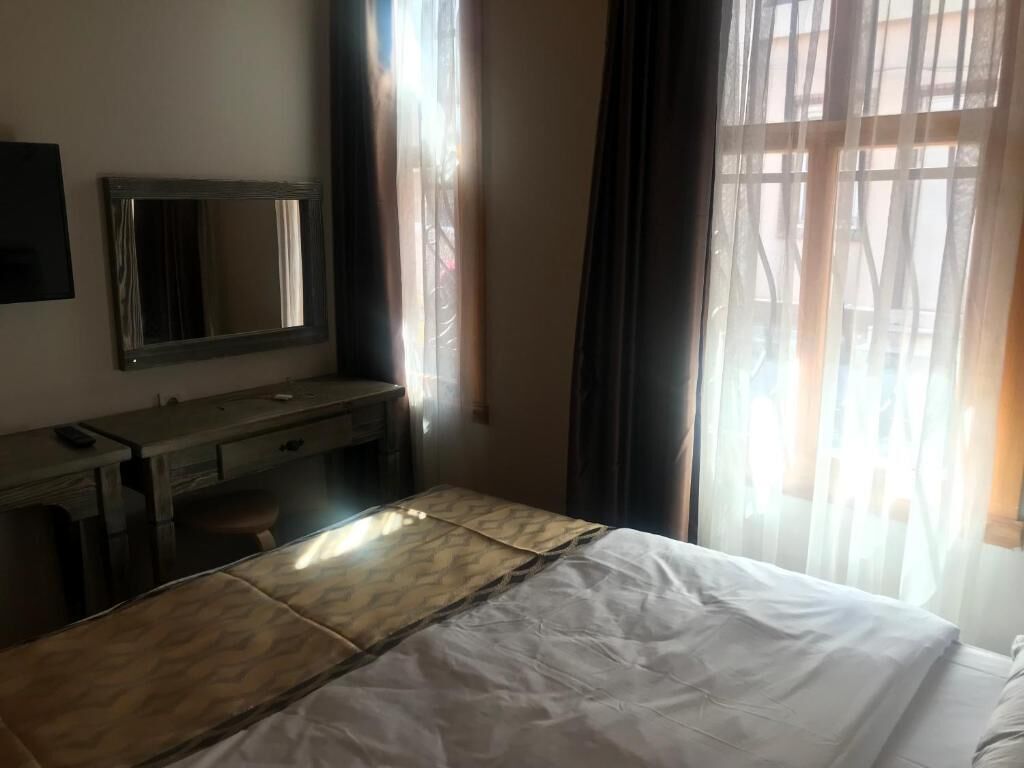 A hotel room with a bed, a desk, a chair, a mirror, and sheer curtains.