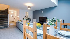 A modern living room with a staircase, a dining area with wooden chairs and a glass table, and a TV on the wall. - 3br-sleeps6-garden-bbq-freeparking-pets (Salford)