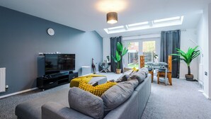 A modern living room with a grey sofa, a flat-screen TV, a dining table with chairs, and a large window with curtains. - 3br-sleeps6-garden-bbq-freeparking-pets (Salford)