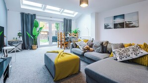 A modern living room with a grey sofa, yellow throw blanket, and a dog. There is a dining table with chairs, a large plant, and a view of the outdoors through a glass door. - 3br-sleeps6-garden-bbq-freeparking-pets (Salford)