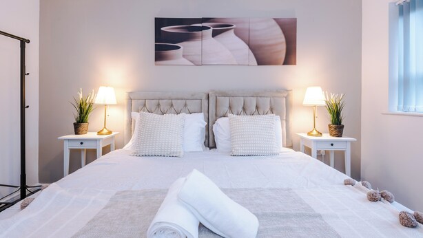 A neatly made bed with a tufted headboard, two bedside tables with lamps and potted plants, and a large abstract painting above the bed. - 3br-sleeps6-garden-bbq-freeparking-pets (Salford)