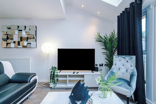 Ein modernes Wohnzimmer mit einem Flachbildfernseher, einem schwarzen Ledersofa, einem weißen Couchtisch und einer Zimmerpflanze.