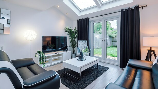 A modern living room with a flat-screen TV, a white coffee table, black leather sofas, a potted plant, and a large window with curtains. - 3br-walktocitycentre-garden-freeparking (Salford)