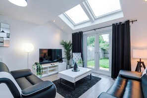 Ein modernes Wohnzimmer mit einem Flachbildfernseher, einem weißen Couchtisch, schwarzen Ledersofas, einer Topfpflanze und einem großen Fenster mit Vorhängen.